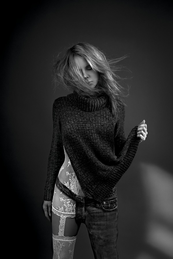 Black and white portrait of a blonde woman wearing both white lace lingerie and a wool sweater with a one-legged jean her hair floating in the wind by Artmood Visualz